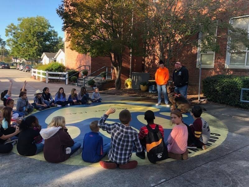 Trainer presenting dog to elementary school students seated in outdoor courtyard